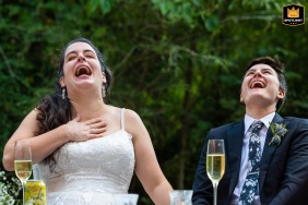 In the backyard of a home in Atlanta, Georgia, the couple sits at the head table during wedding speeches, laughing together with their heads tilted back, capturing their shared joy and the lively, relaxed atmosphere of the celebration.